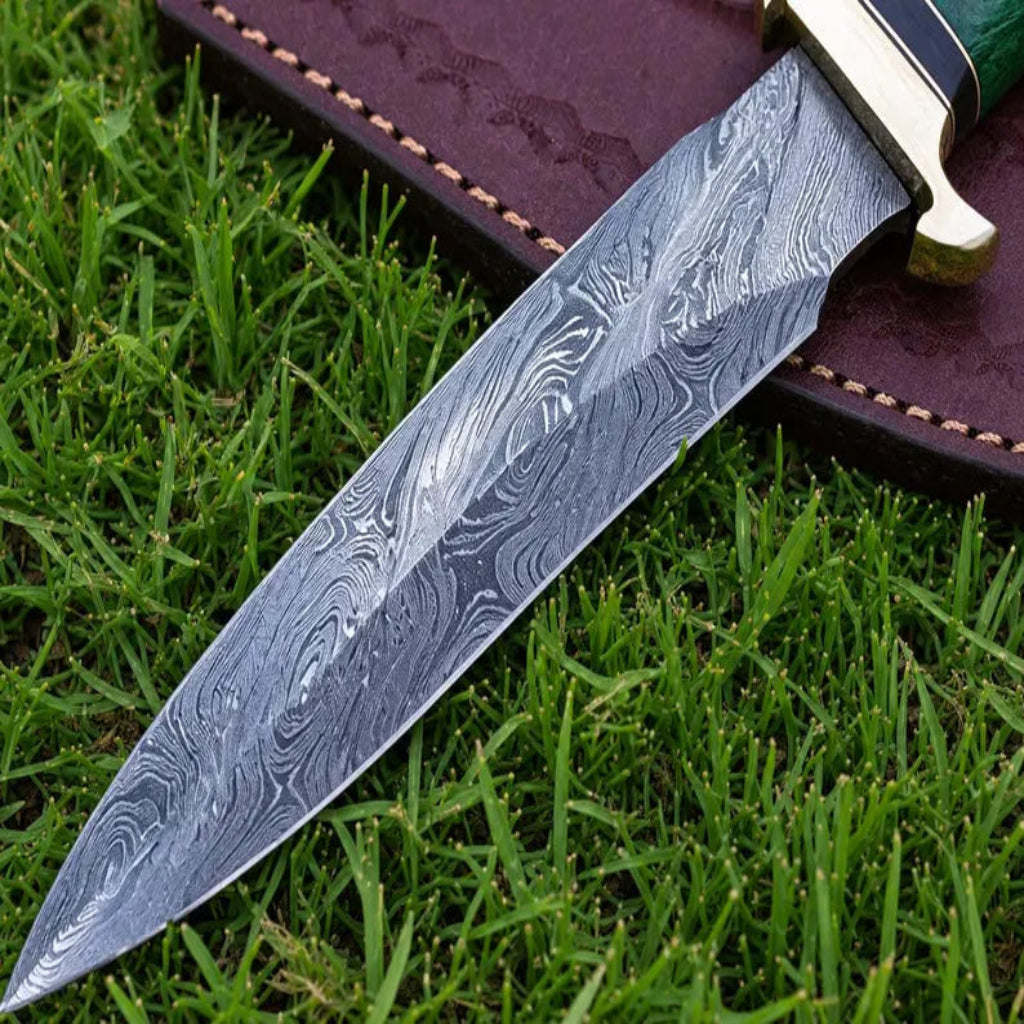 A close-up of a Damascus steel knife with a wavy pattern blade, resting on green grass next to a brown leather sheath.