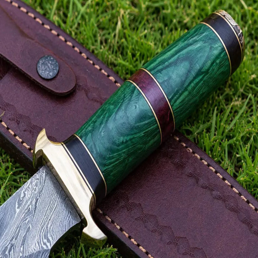 A close-up of a custom fixed-blade knife with a green wood-grain handle and Damascus steel blade, resting on its dark brown leather sheath over a green grass background.